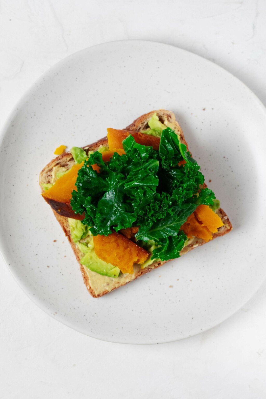 An image of a slice of avocado toast, topped with winter squash and kale.