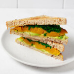 An angled image of a vegan sandwich made with layers of roasted kabocha squash, avocado, and kale. The sandwich is served on a small, round white plate.
