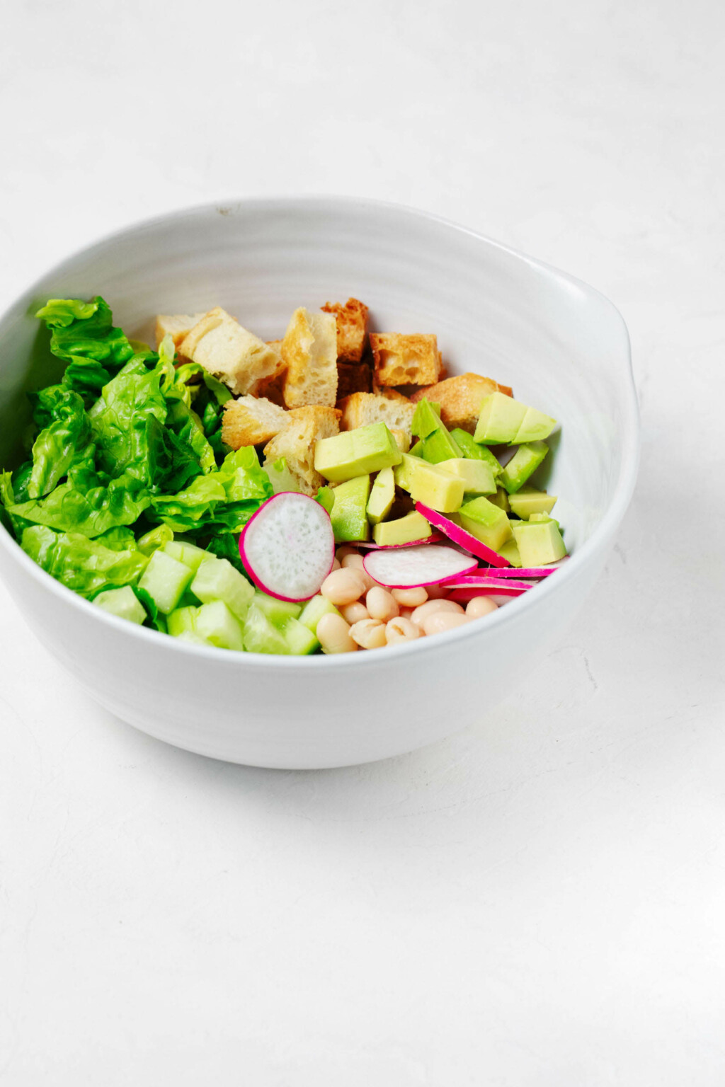 A large, white serving bowl is filled with cubed bread, radishes, white beans, and greens.