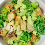 An overhead image of a white bowl, which is filled with lettuces, white beans, radishes, avocado, and a creamy green dressing.