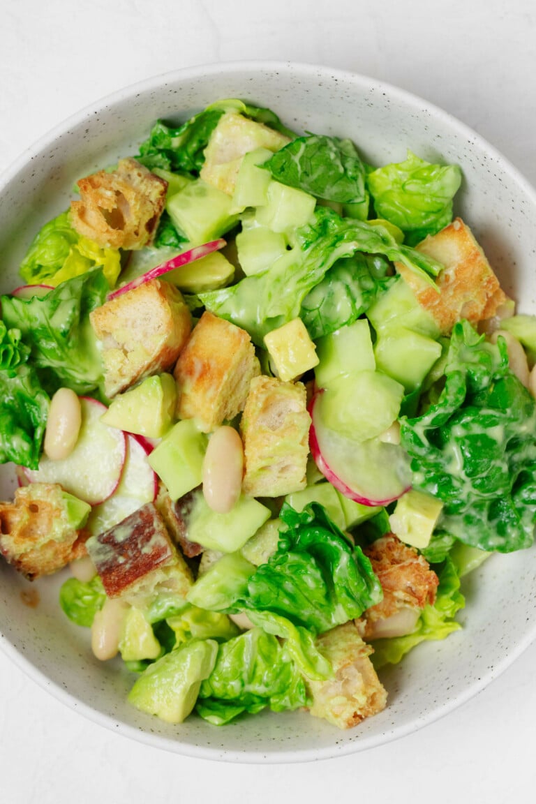 An overhead image of a white bowl, which is filled with lettuces, white beans, radishes, avocado, and a creamy green dressing.