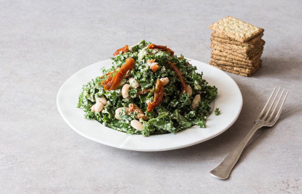 A white, round serving plate has been piled high with a kale white bean Caesar salad. A small stack of crackers is visible behind it.