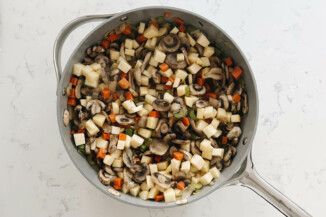 A large skillet is resting on a white surface. It holds celery, carrots, onions, mushrooms, and chopped rutabaga.