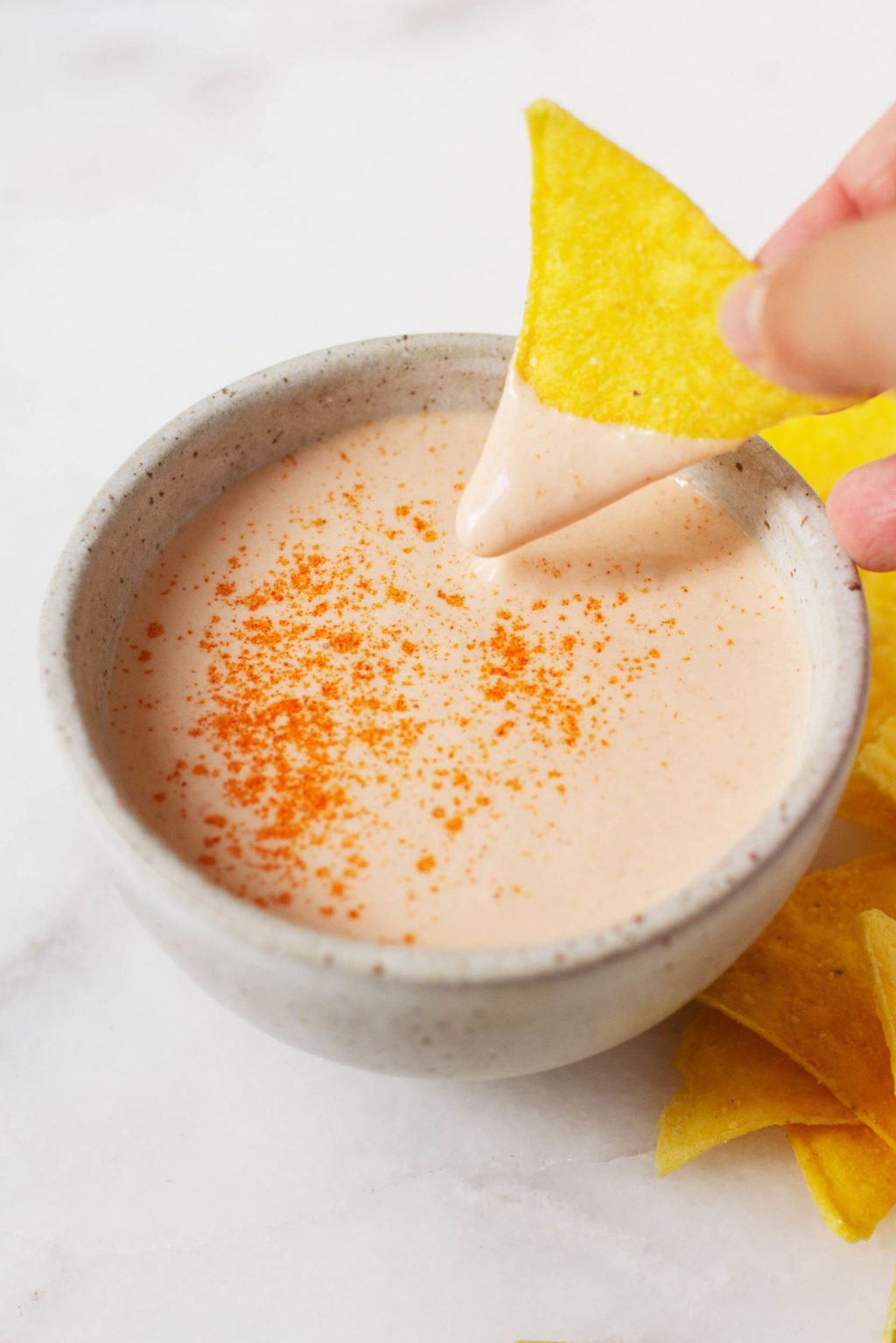 A triangular corn chip is being dipped into a small pinch bowl of a creamy, plant-based sauce.