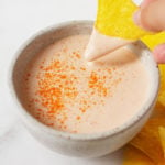A triangular corn chip is being dipped into a small pinch bowl of a creamy, plant-based sauce.