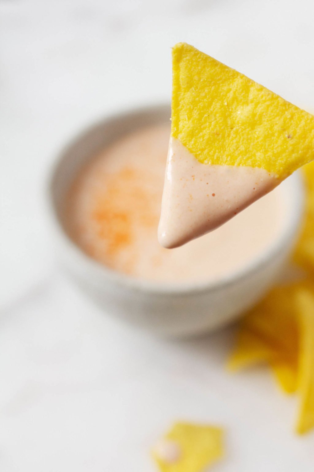 A single corn chip has been dipped in a creamy vegan queso sauce. The bowl of queso is pictured in the background, along with extra chips.