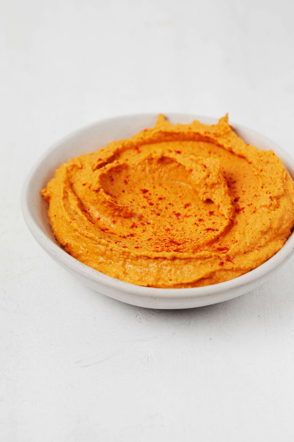 A vegan sweet potato hummus has been topped with a bit of paprika. It's resting in a small, white bowl.