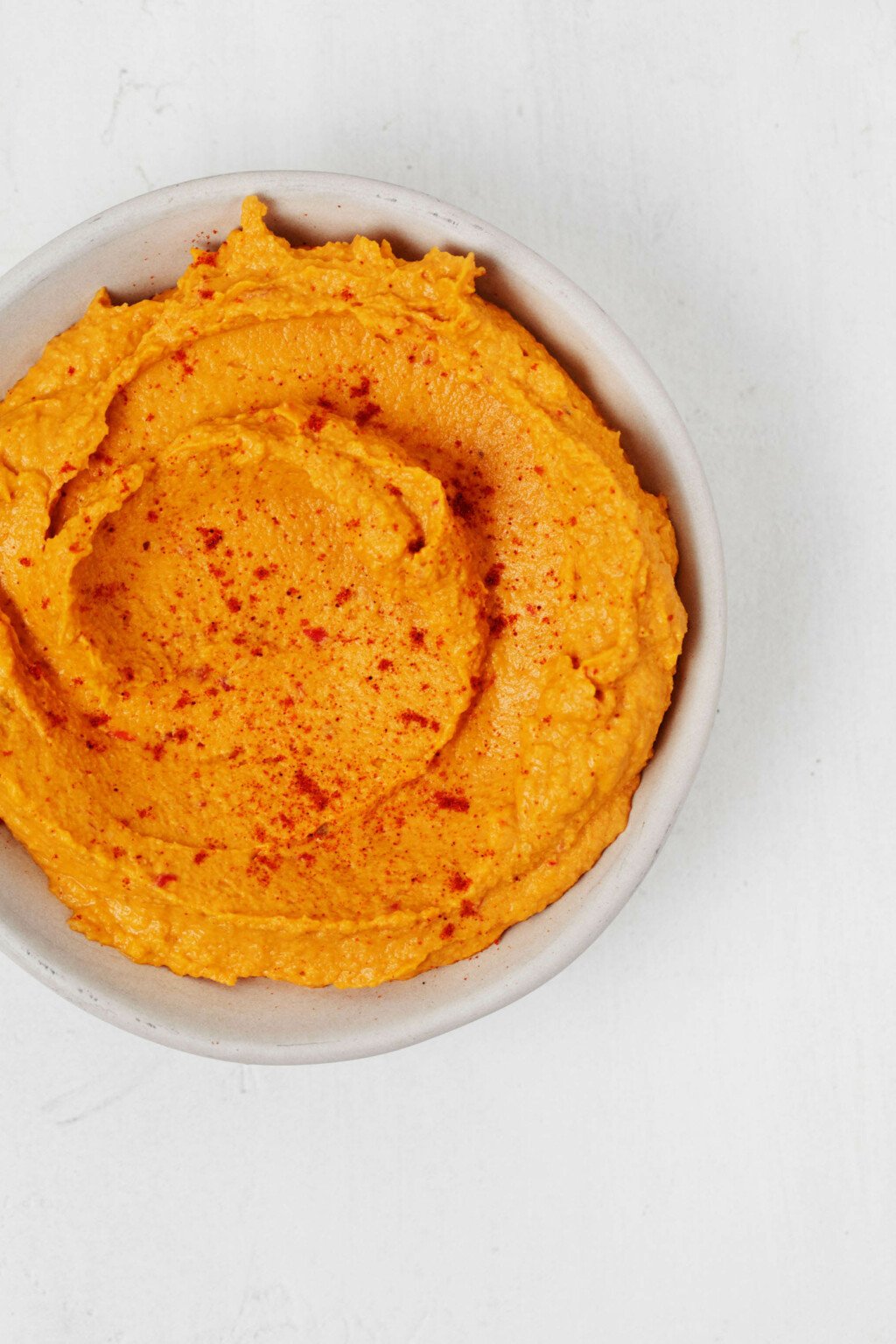 A swirled, vegan dip, which is made with sweet potatoes, is in a white bowl that rests on a white surface.