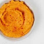 A swirled, vegan dip, which is made with sweet potatoes, is in a white bowl that rests on a white surface.