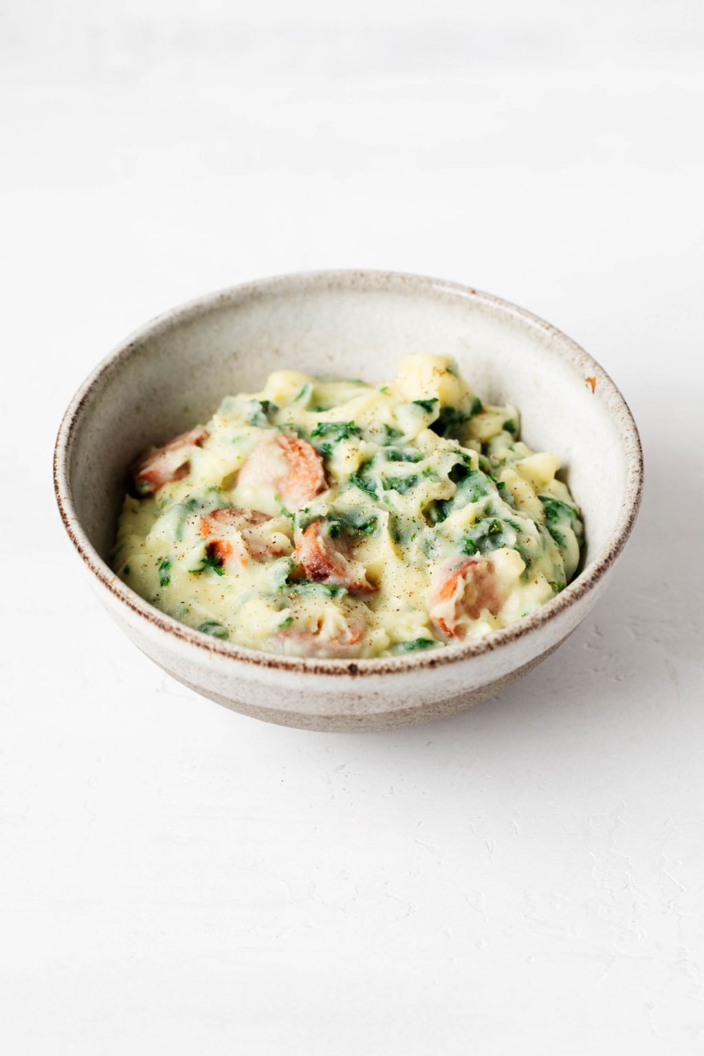 An angled photograph of a ceramic bowl holding a vegan kale colcannon recipe. It rests on a white surface.