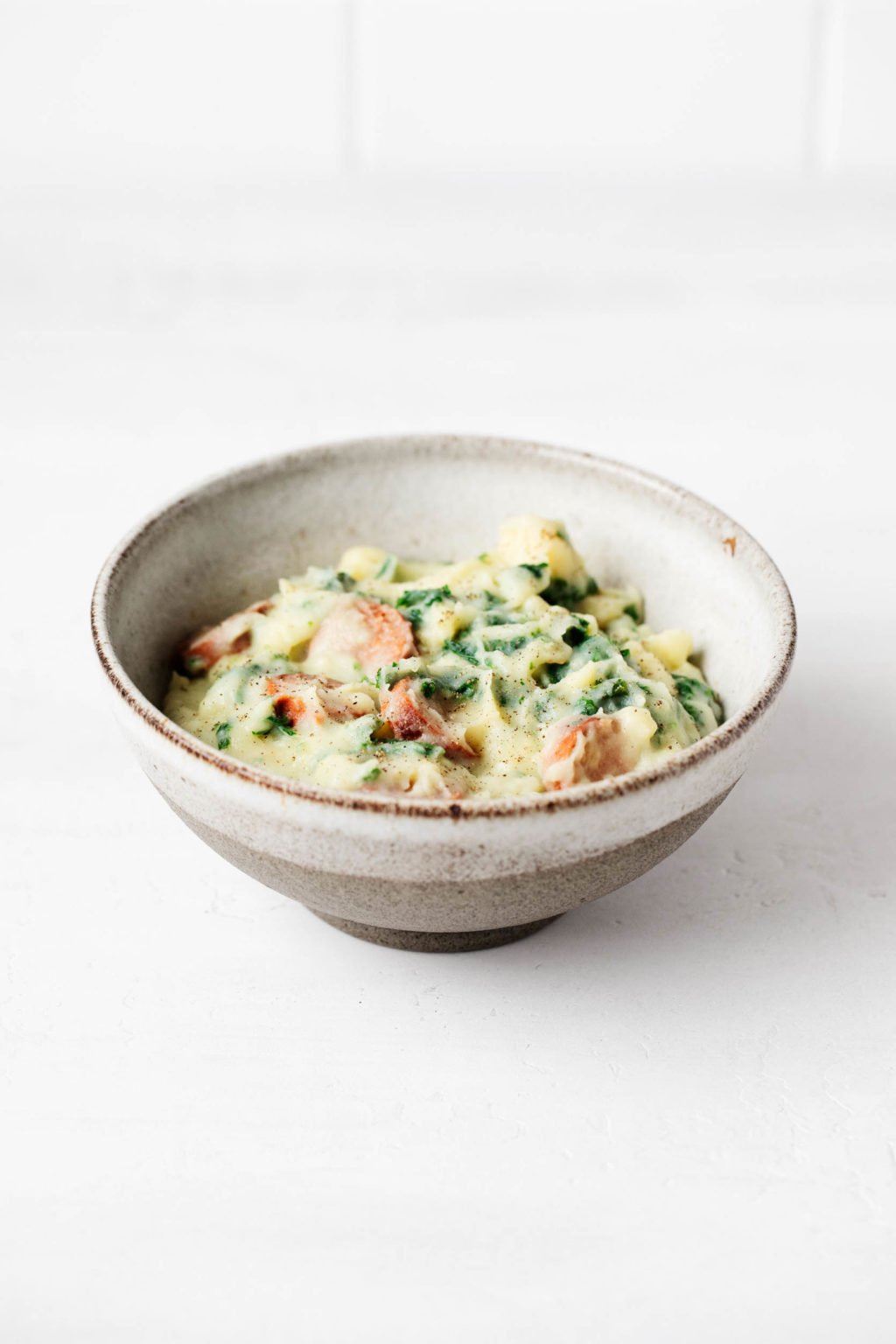 A white and gray ceramic, stoneware bowl is resting on a white surface. It contains a mashed potato and vegetable mixture.