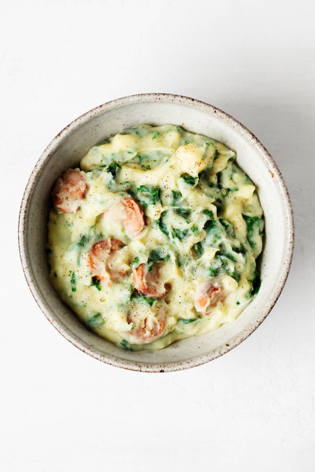 A round, white ceramic bowl has been filled with mashed potatoes, green vegetables, and sausage slices.