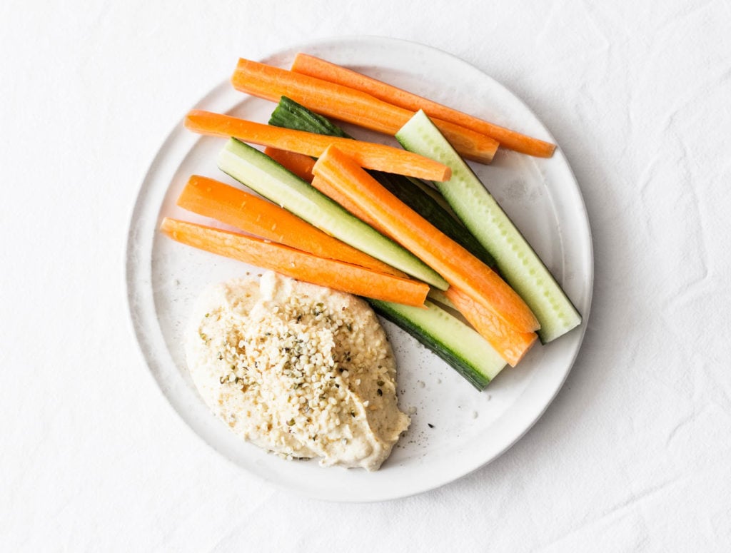 An overhead image of a snack plate with hemp seed hummus, carrots, and raw cucumbers.