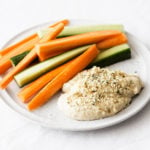 A round, white plate is covered in ingredients for a snack, including a vegan hemp hummus and raw crudites.