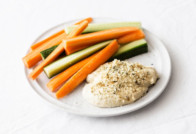 A round, white plate is covered in ingredients for a snack, including a vegan hemp hummus and raw crudites.