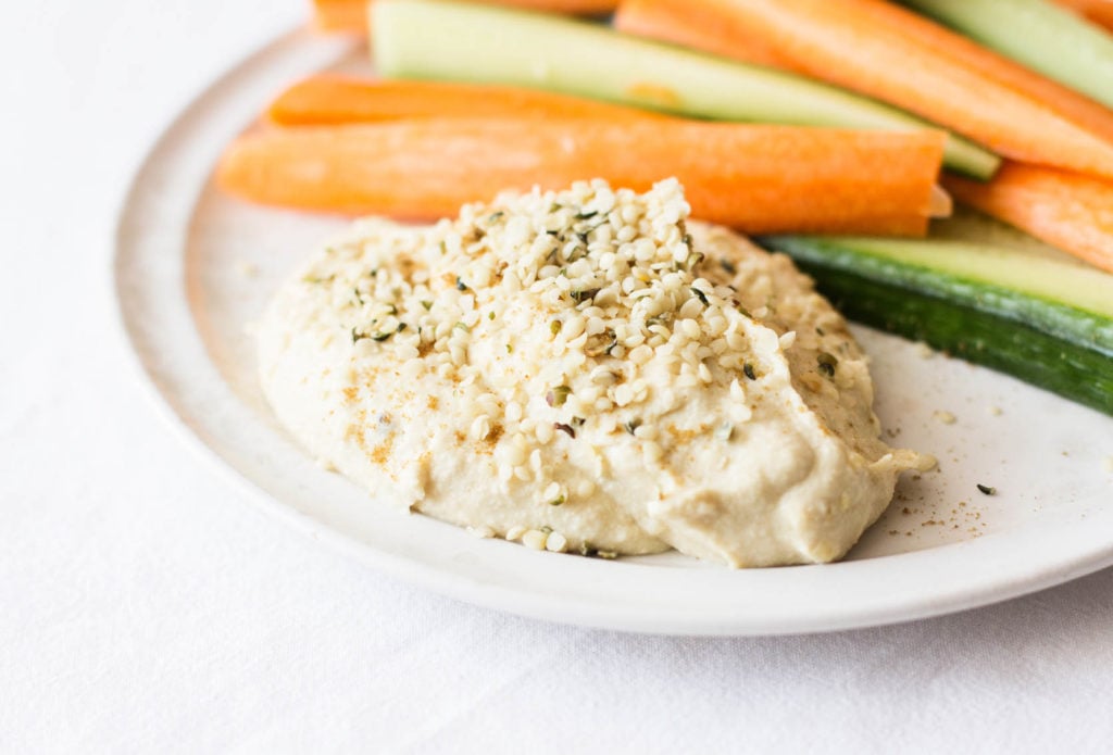 A zoomed in photograph of hummus on a small white serving plate. The hummus has been sprinkled with shelled hemp seeds.