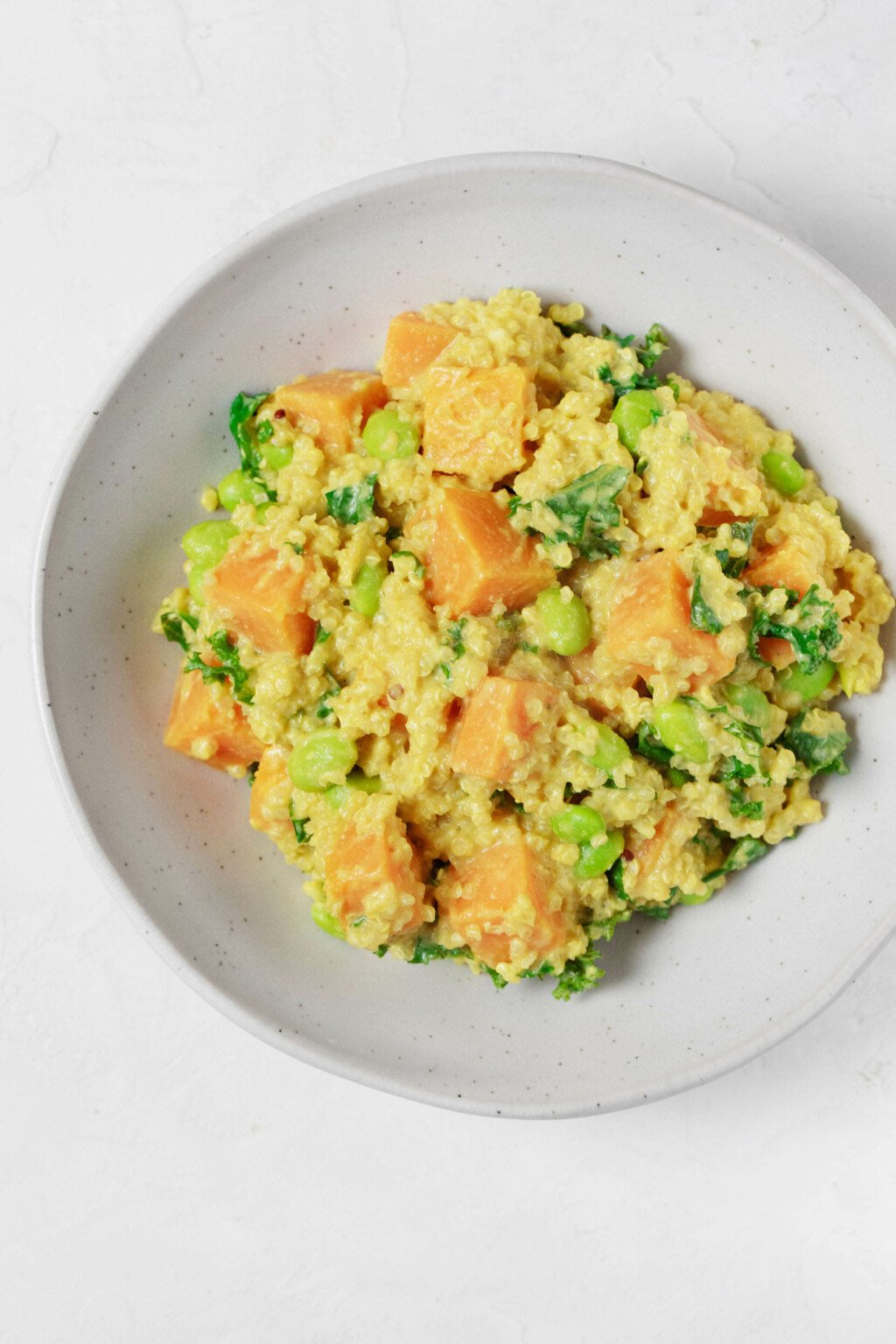 An overhead image of a round white bowl, which has been filled with a golden-hued mixture of quinoa, edamame, and butternut squash.