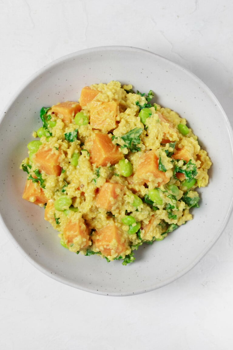 An overhead image of a round white bowl, which has been filled with a golden-hued mixture of quinoa, edamame, and butternut squash.