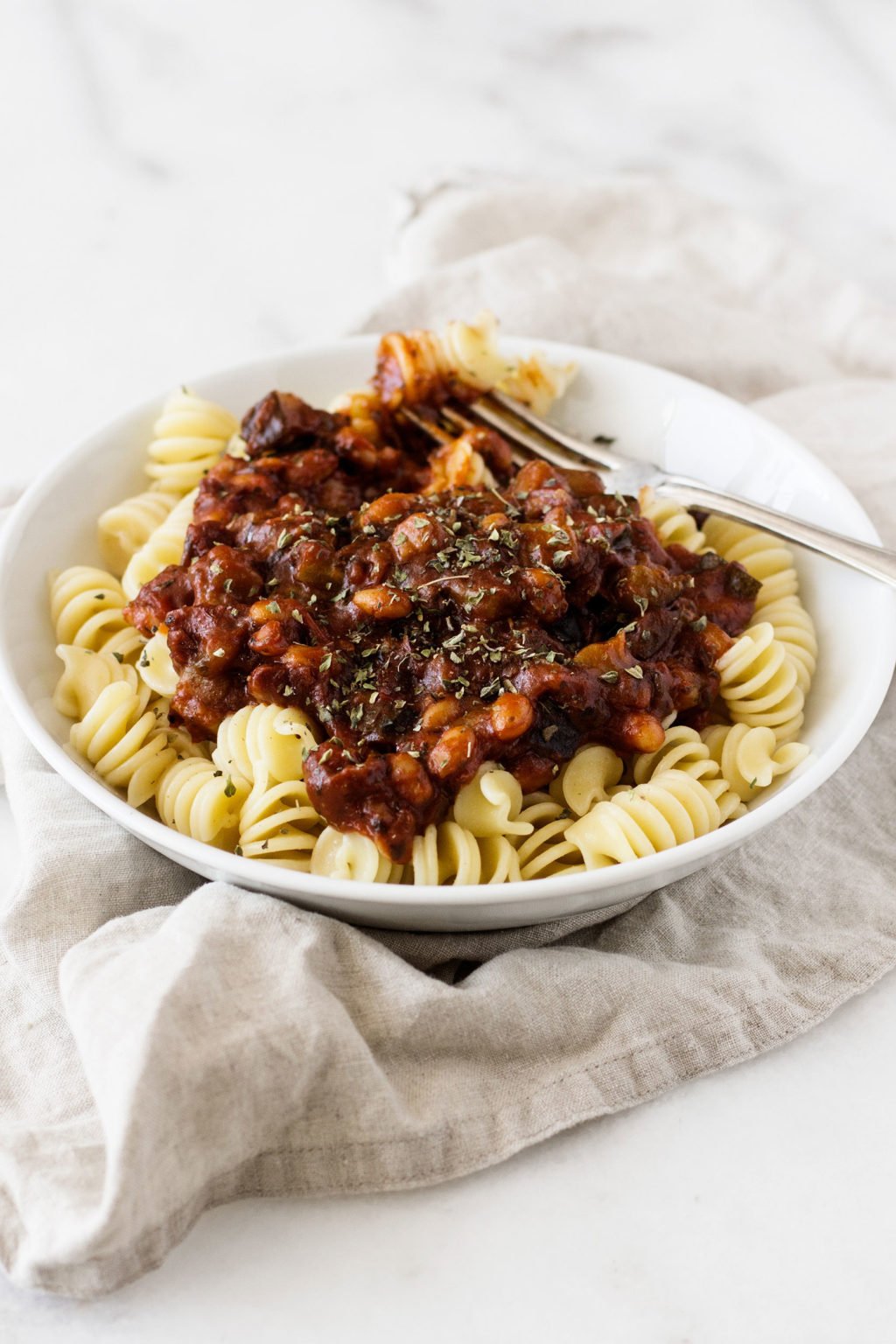 A beige, linen napkin is lying underneath a white bowl of pasta, beans, and vegetables.