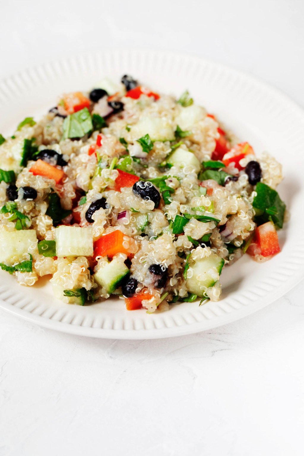 An angled image of a colorful quinoa black bean salad, which has been piled onto a fluted, white ceramic plate. The plate is on a white surface.