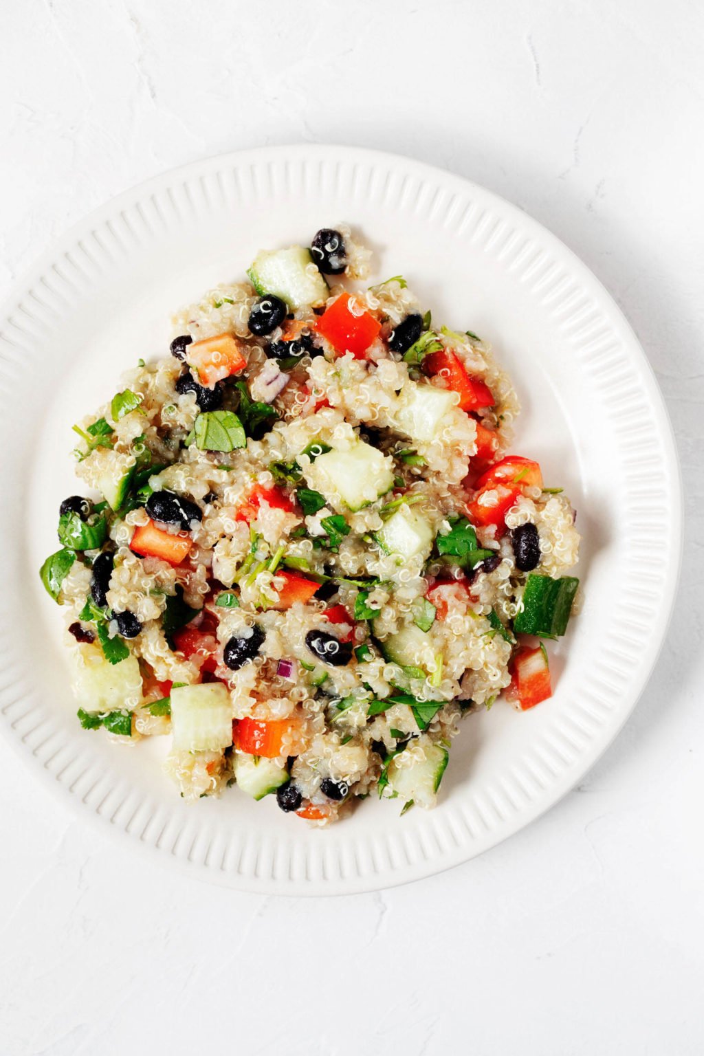 A white salad plate with a fluted rim has been piled with a plant-based grain salad. The salad is full of colorful vegetables and fresh herbs.