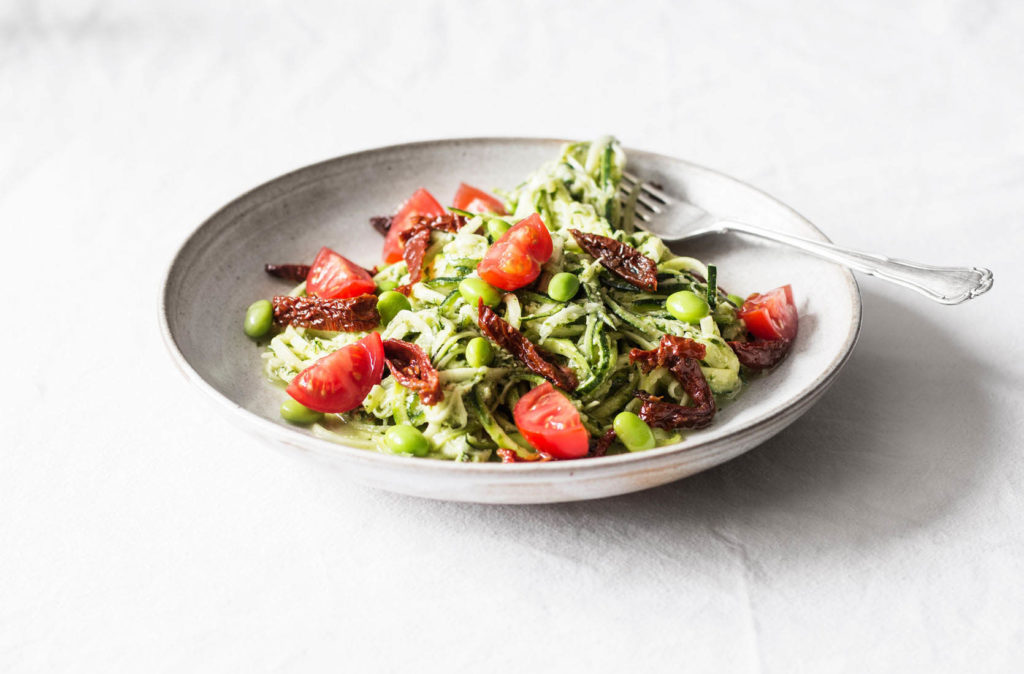 An angled photograph of zucchini noodles dressed in a hemp seed pesto. The noodles are tossed with edamame and cherry tomatoes.