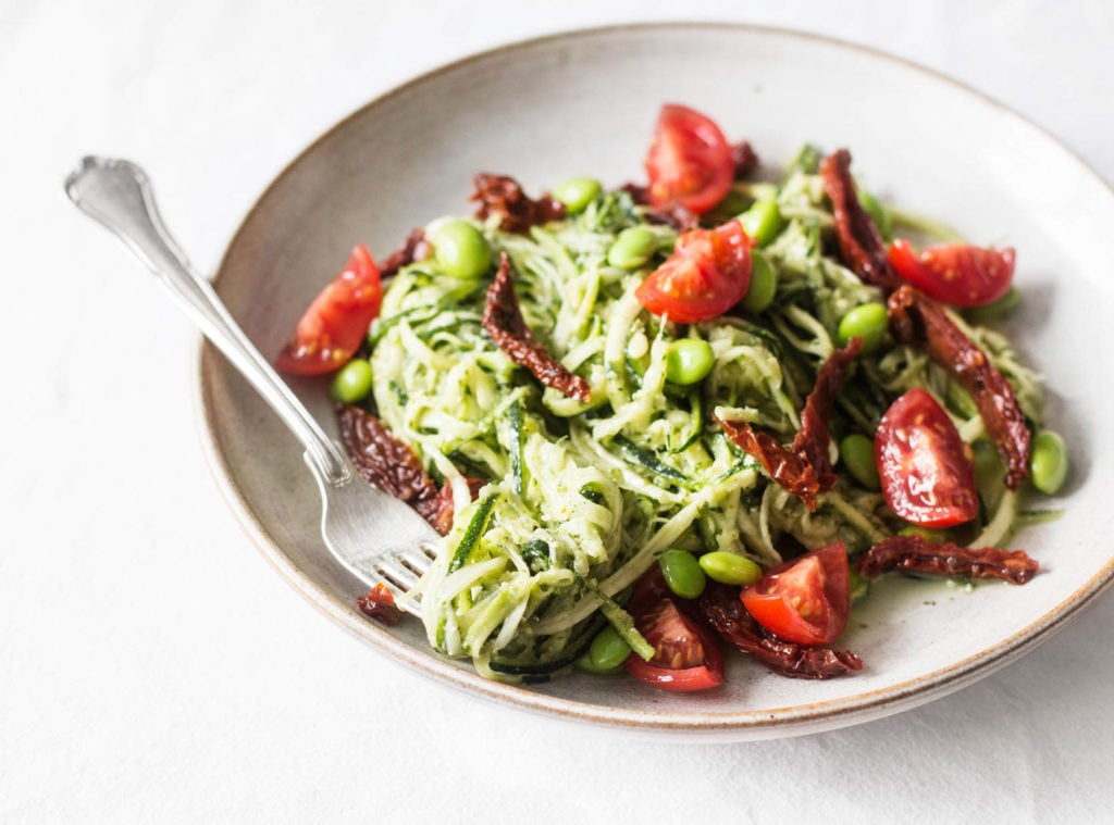 A white, ceramic bowl with a light gray rim is filled with summery zucchini noodles, tomato, and pesto.