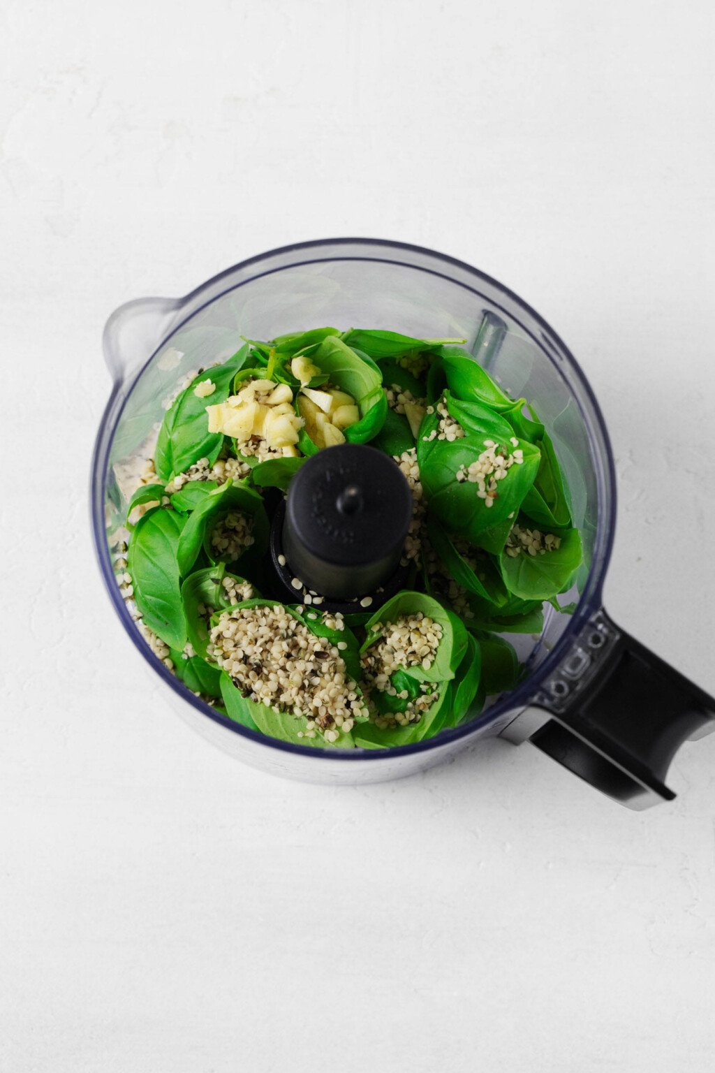 Basil leaves, nutritional yeast, and salt are pictured in the bowl of a food processor fitted with the S blade.