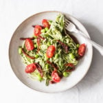 A white and light gray ceramic bowl is filled with zucchini noodles dressed in a vegan hemp seed pesto, edamame, and cherry tomatoes.