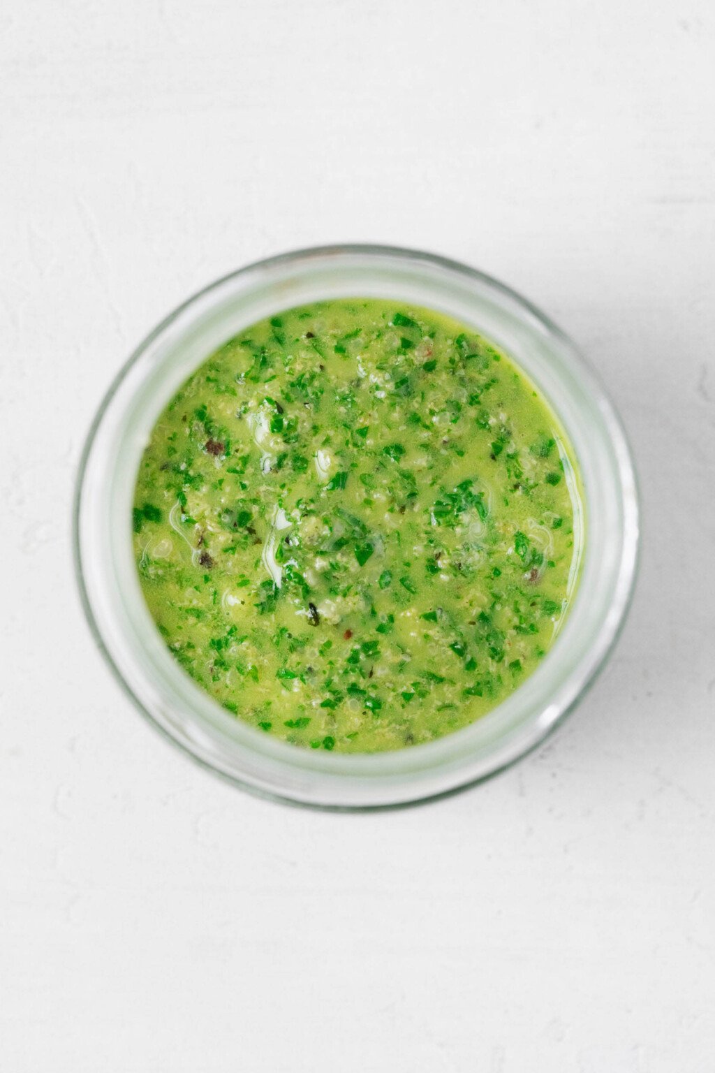 A green vegan pesto sauce with hemp seeds has been placed into a round, clear jar, pictured from above.