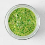 A green vegan pesto sauce with hemp seeds has been placed into a round, clear jar, pictured from above.