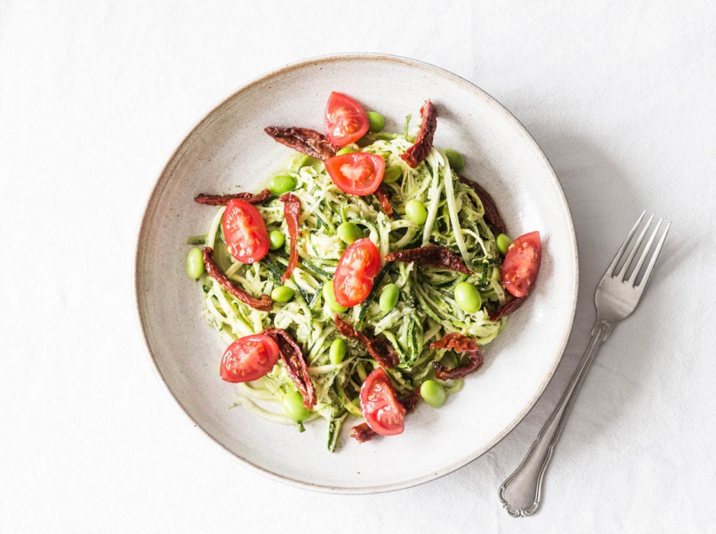 A white bowl is filled with zucchini noodles, vegetables, and a green herb sauce. It's placed on a white tablecloth.