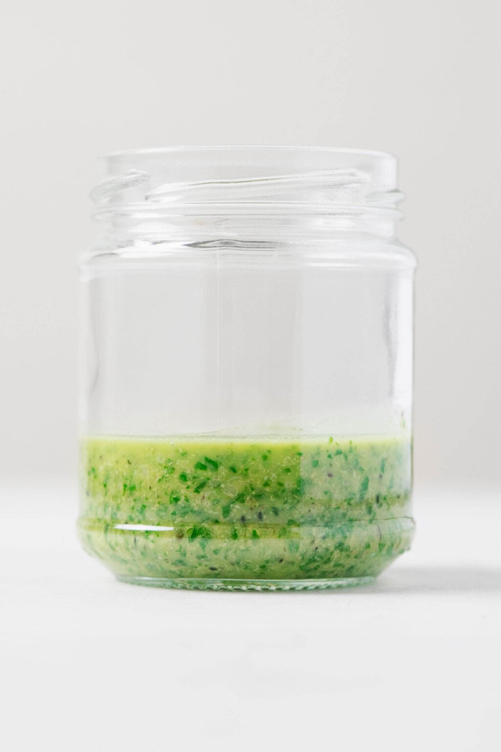 A green vegan pesto sauce with hemp seeds has been placed into a round, clear jar, pictured resting on a white surface.