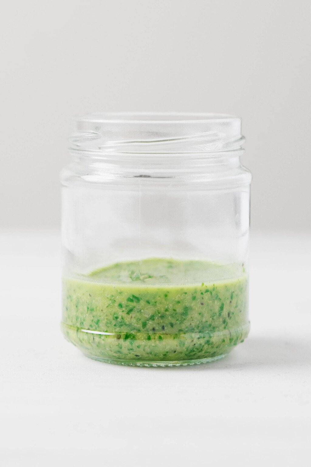 A green vegan pesto sauce with hemp seeds has been placed into a round, clear jar, pictured resting on a white surface.