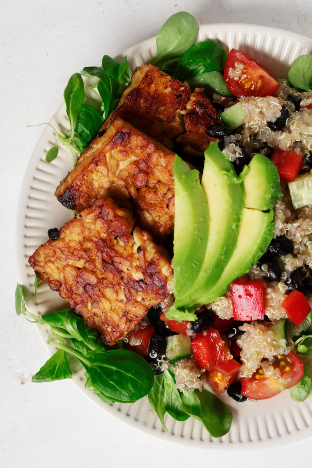 Seared, square slices of tempeh are arranged with avocado and vegetables on a salad plate.
