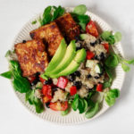 An overhead image of a rimmed salad plate, which is covered in greens, quinoa, and seared tempeh slices.