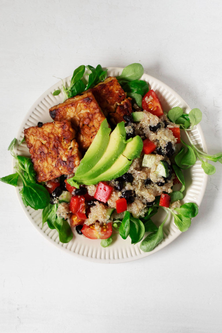 An overhead image of a rimmed salad plate, which is covered in greens, quinoa, and seared tempeh slices.