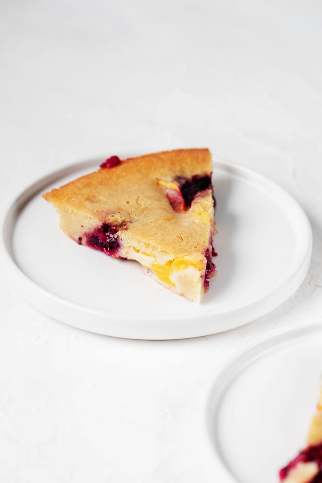 An angled photograph of a slice of vanilla cake with berries. It's served on a round dessert plate, which is resting on a white surface.