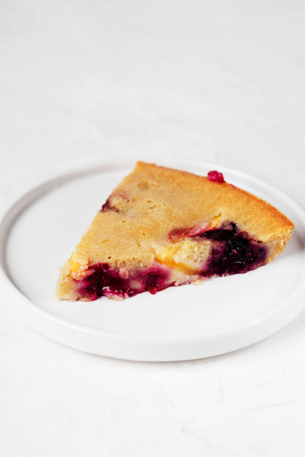 An angled photograph of a slice of vegan peach blackberry skillet cake, which is resting on a white dessert plate.