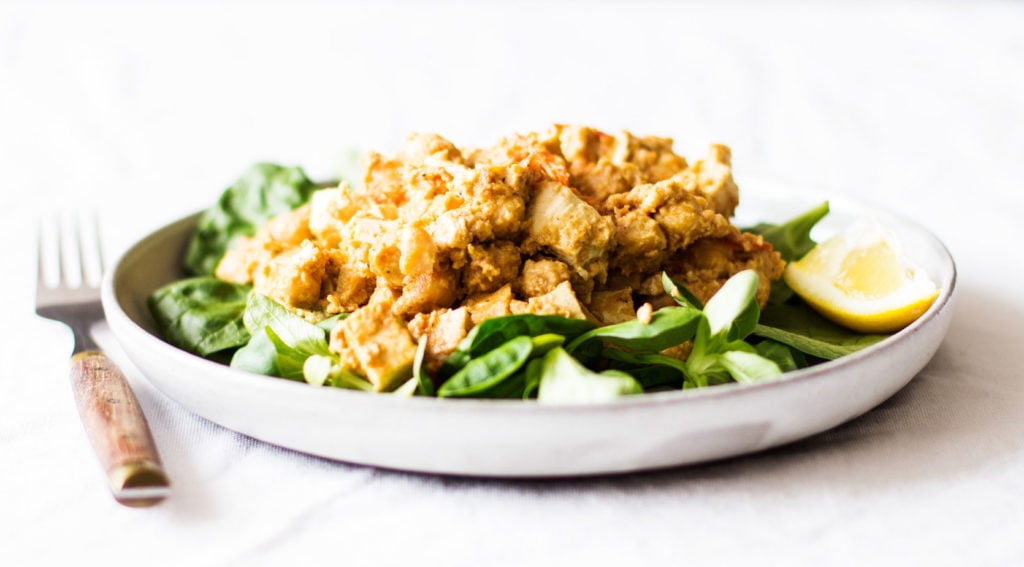 A round, white plate with a small rim has been covered in a tofu tahini scramble, baby spinach, and a wedge of fresh lemon. A fork with a wooden handle rests to the side of the plate.