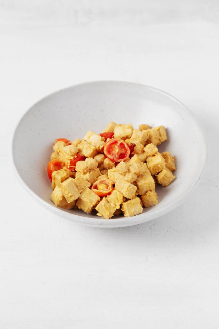 An asymmetrical white bowl rests on a white surface. It holds a tofu tahini scramble that's made with cubed tofu, chickpeas, and halved grape tomatoes.