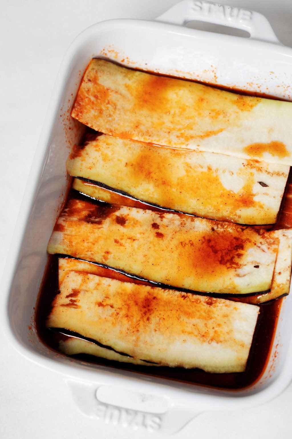 Very thin slices of eggplant are in a baking dish. They're being marinated in an orange, brothy liquid.