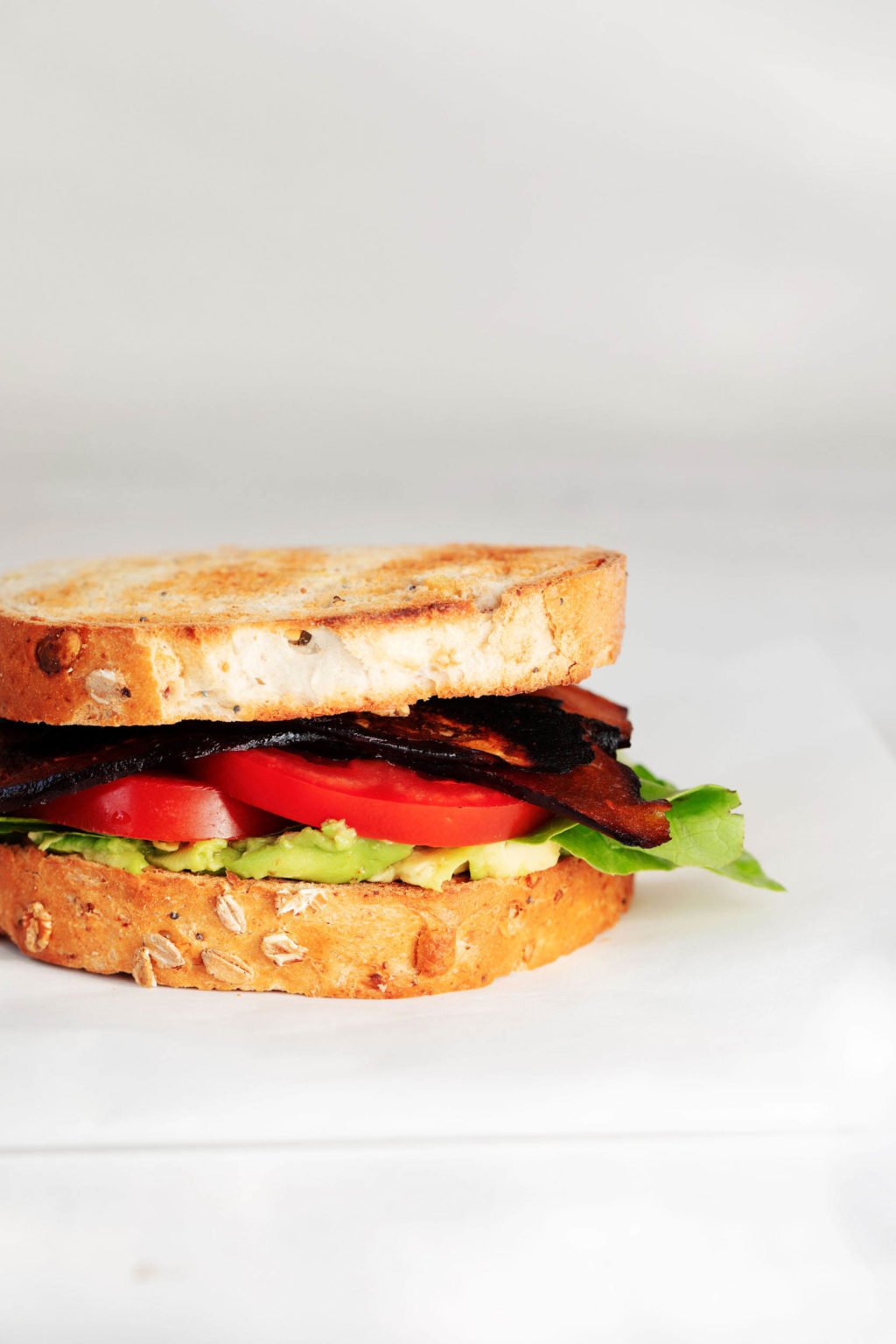 A BLT sandwich is resting on a white surface, against a gray and white background.