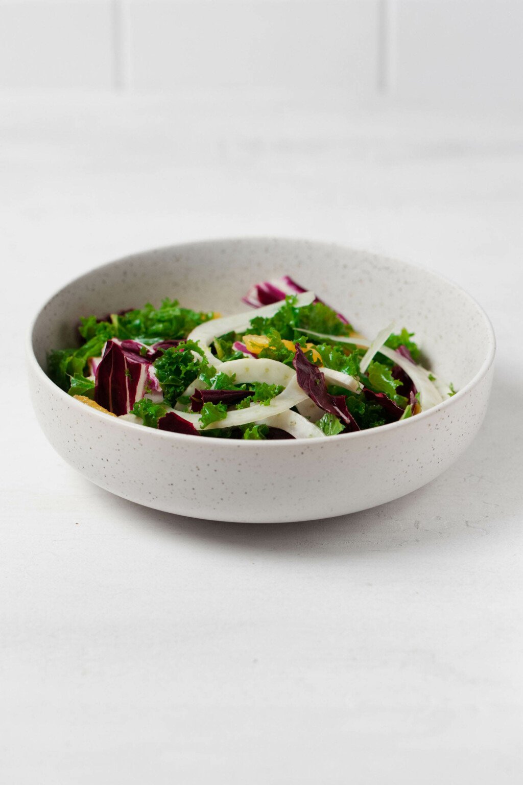 A large, white mixing bowl rests on a white surface. It contains a colorful kale salad.