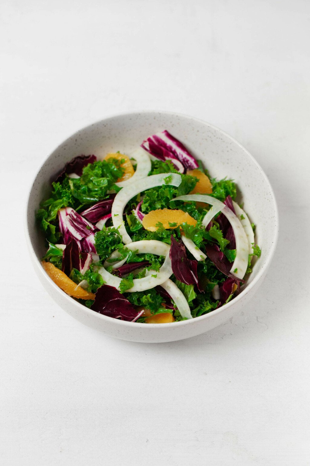 An overhead image of a large white mixing bowl, which is filled with shaved fennel, kale, radicchio, and orange sections.