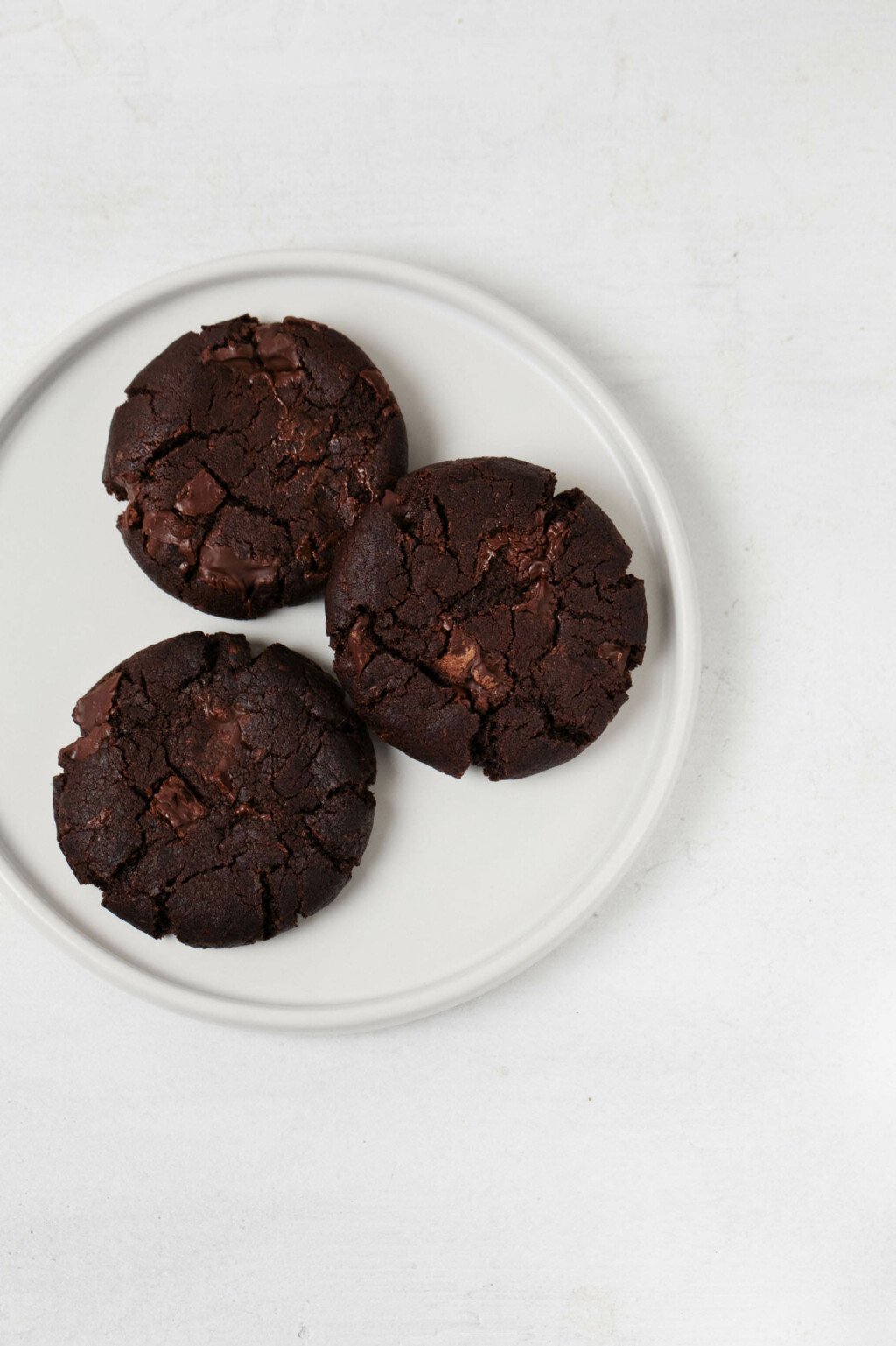 Three round, chewy double chocolate chip cookies are resting on a white ceramic plate. The plate is placed on a white surface.