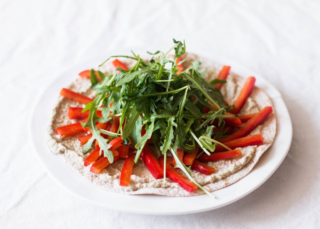 A vegan eggplant tortilla pizza has been laid out on a white tablecloth. It's topped with arugula and bell pepper strips.