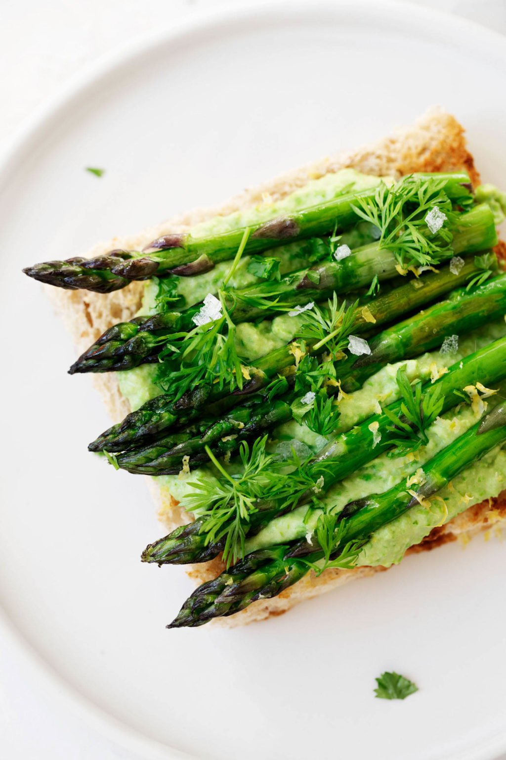 Asparagus spears have been grilled and piled onto toast with a creamy topping. They're garnished with dill, lemon zest, and flaky salt. 