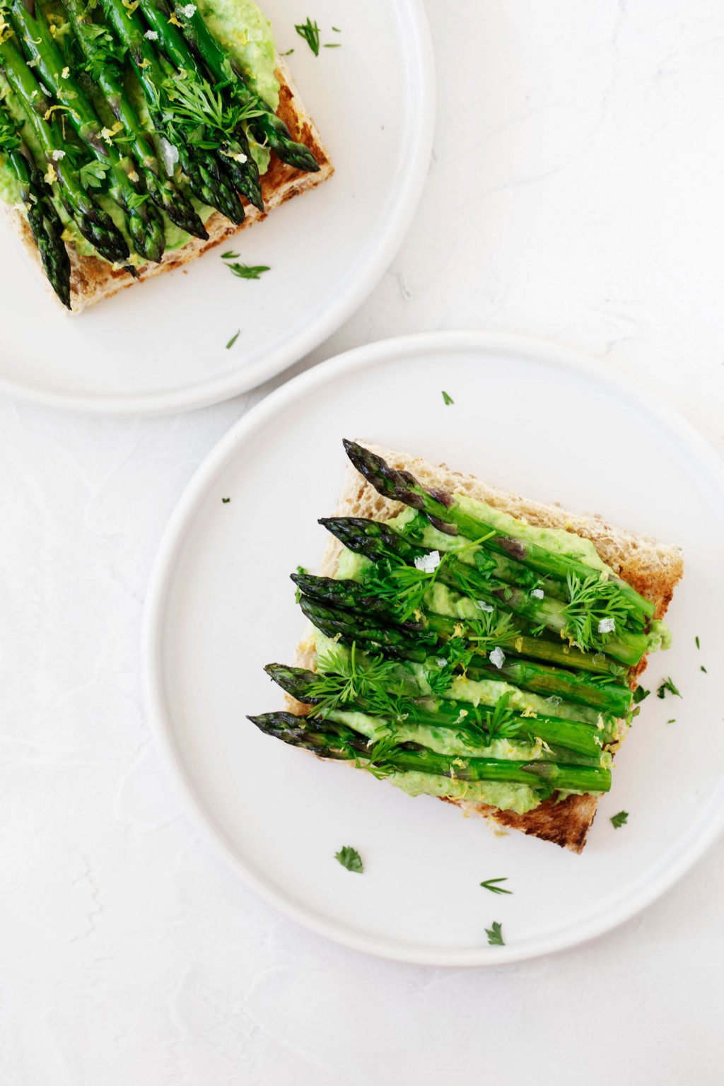 Two round, white plates are laid out on a white surface. They're being used to serve toast with a creamy pea spread and grilled asparagus spears.