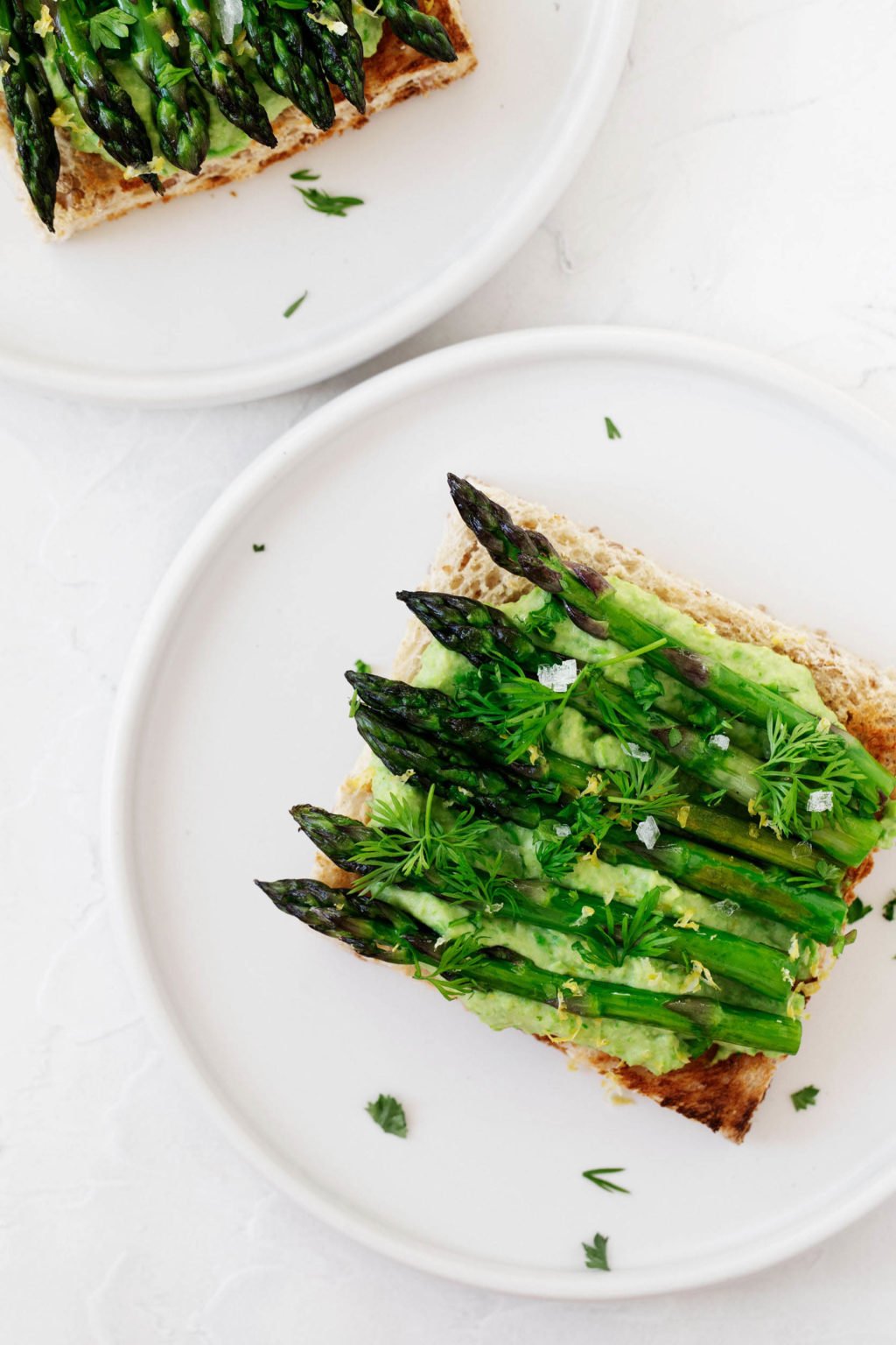 Two small, white appetizer plates carry a vegan green pea hummus tartine, topped with lemon zest and fleur de sel.